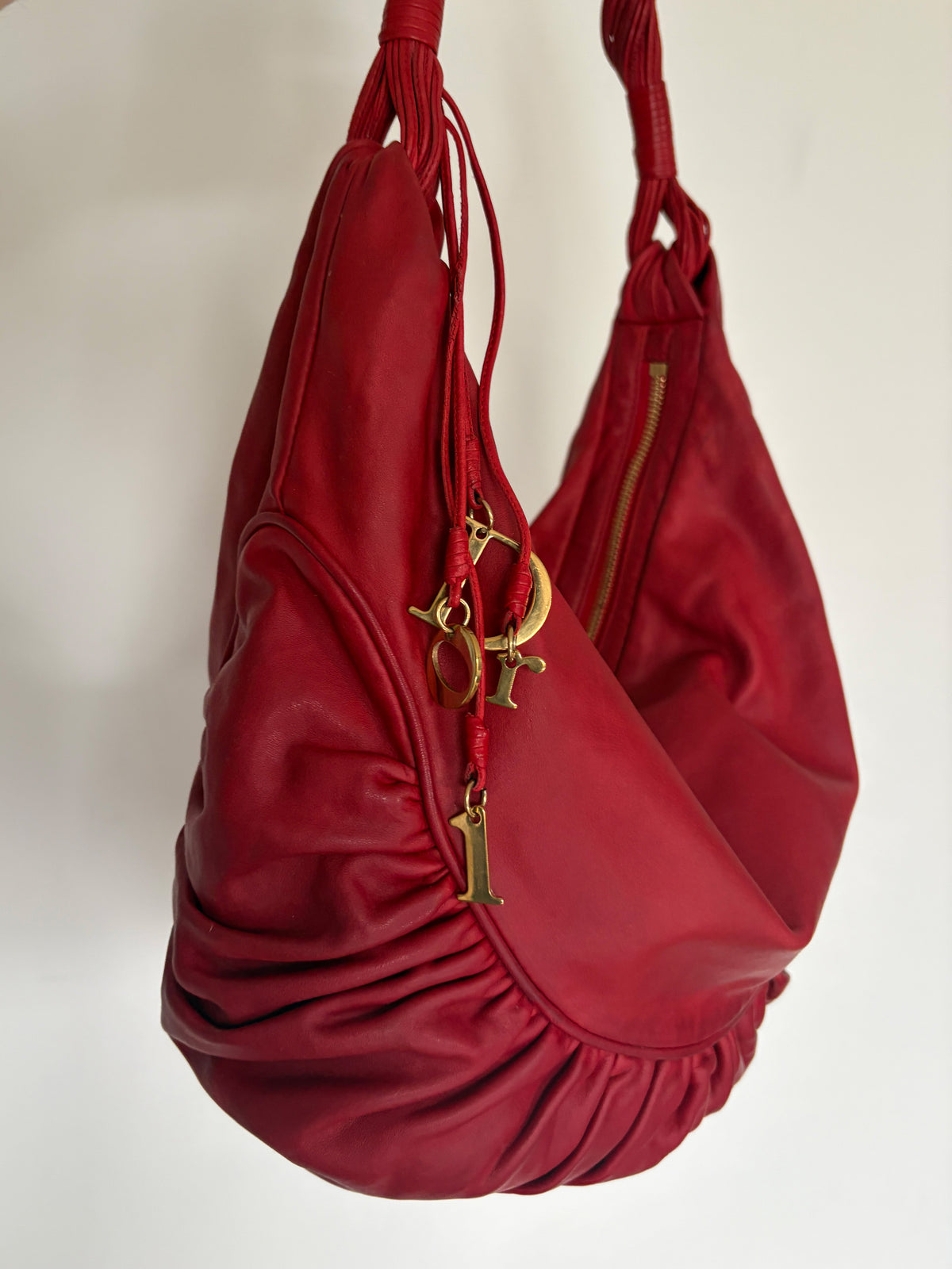 A red leather shoulder bag with gold-tone Christian Dior logo charms hanging from the zipper, shown against a plain off-white background.