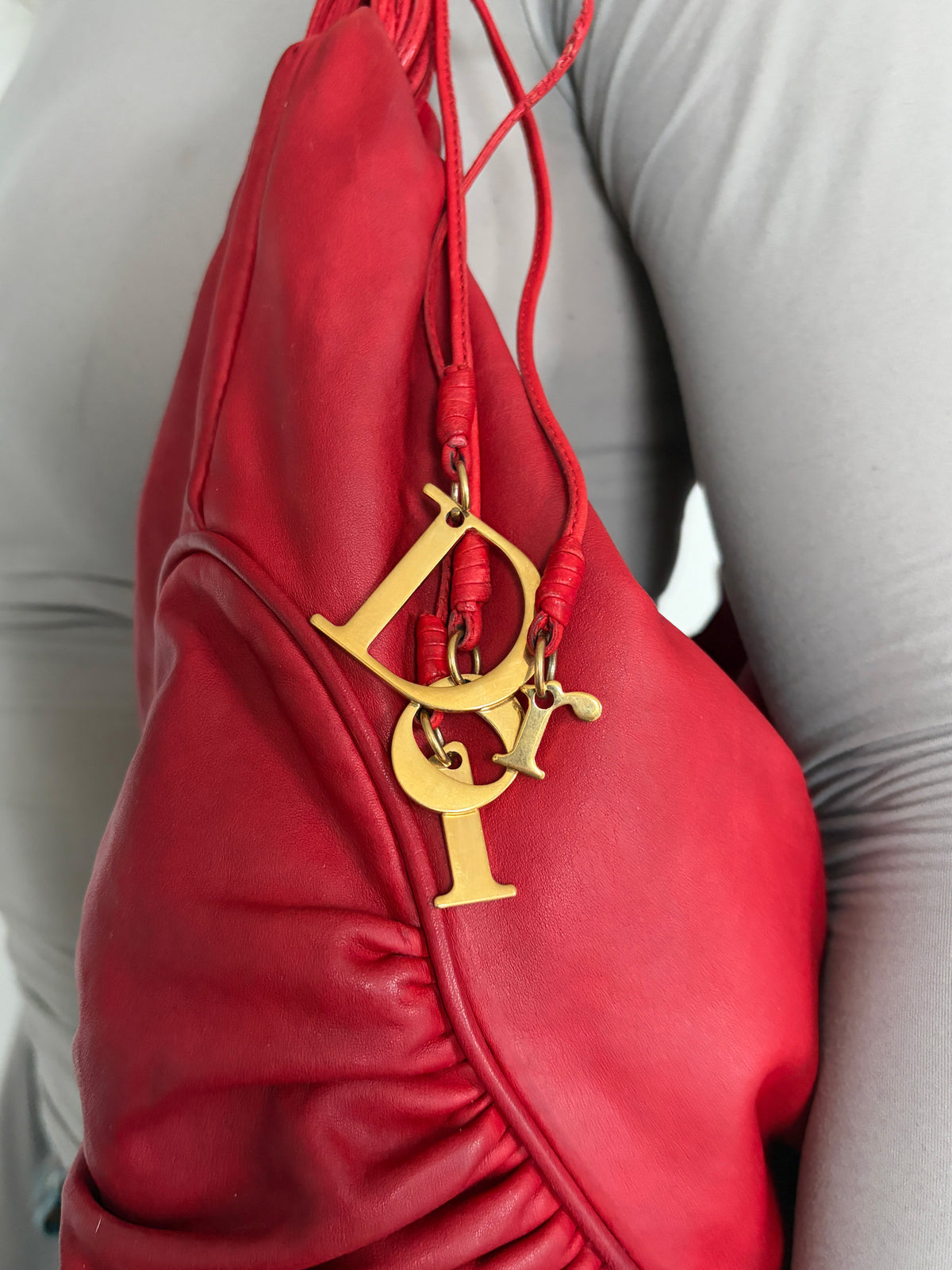 A close-up of a red leather shoulder bag with a gold-tone Christian Dior logo charm, worn by a person in a light gray top.