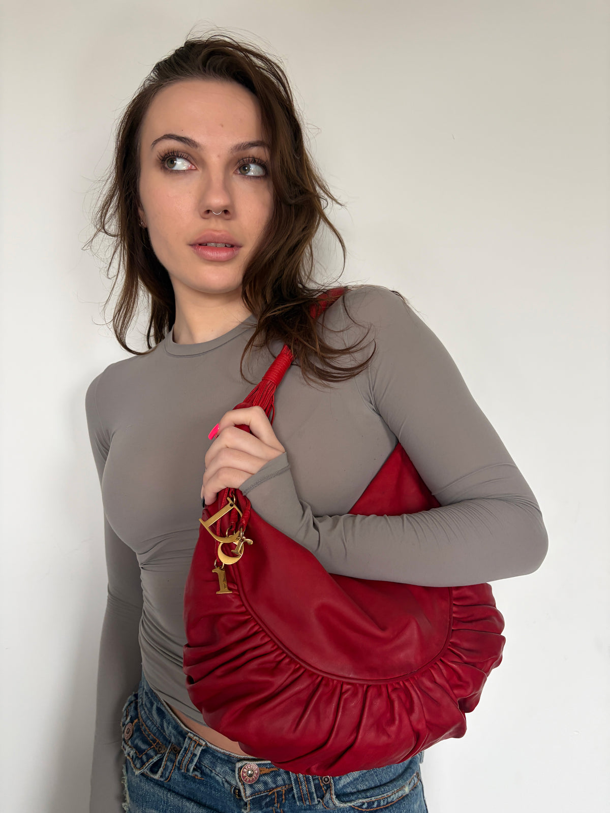 A woman wearing a gray long-sleeve top and blue jeans carries a red crescent-shaped shoulder bag with gold-tone hardware against a plain off-white wall.