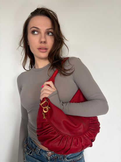 A woman wearing a gray long-sleeve top and blue jeans carries a red crescent-shaped shoulder bag with gold-tone hardware against a plain off-white wall.
